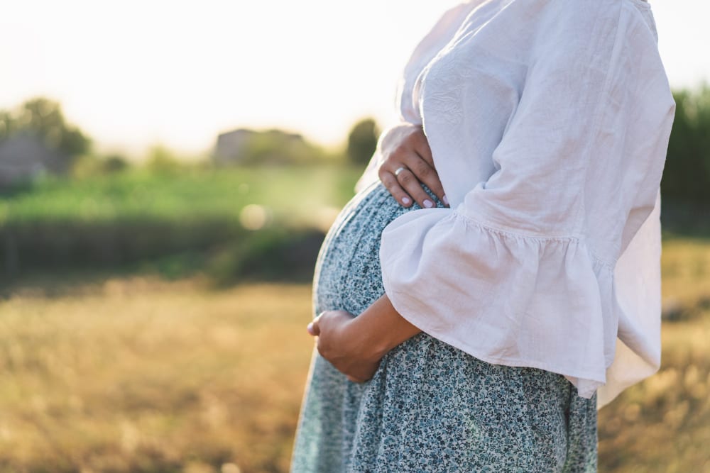 Pregnant woman in a white blouse holding her belly outdoors in warm sunlight