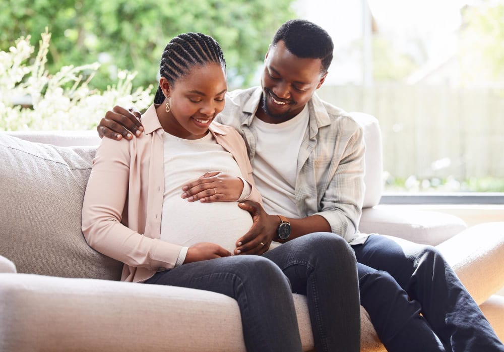 Expectant couple sitting together on a couch, smiling as they hold the pregnant bump